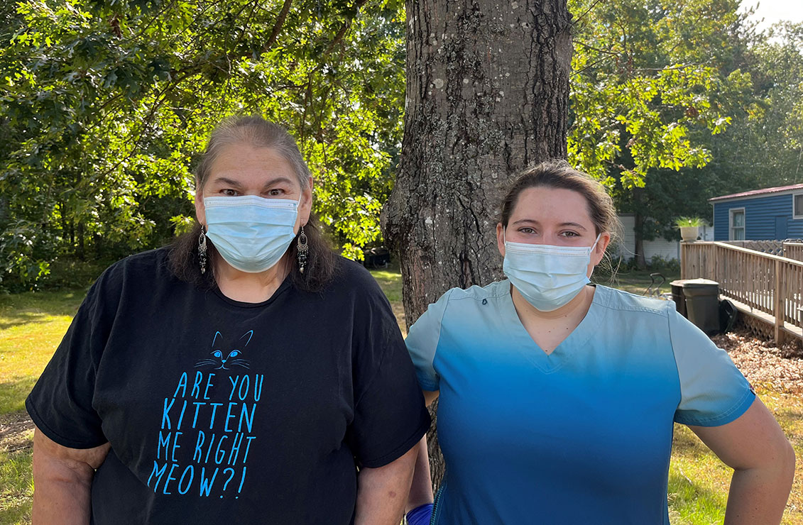 Two women, wearing masks and standing together outside in front of a large leafy tree
